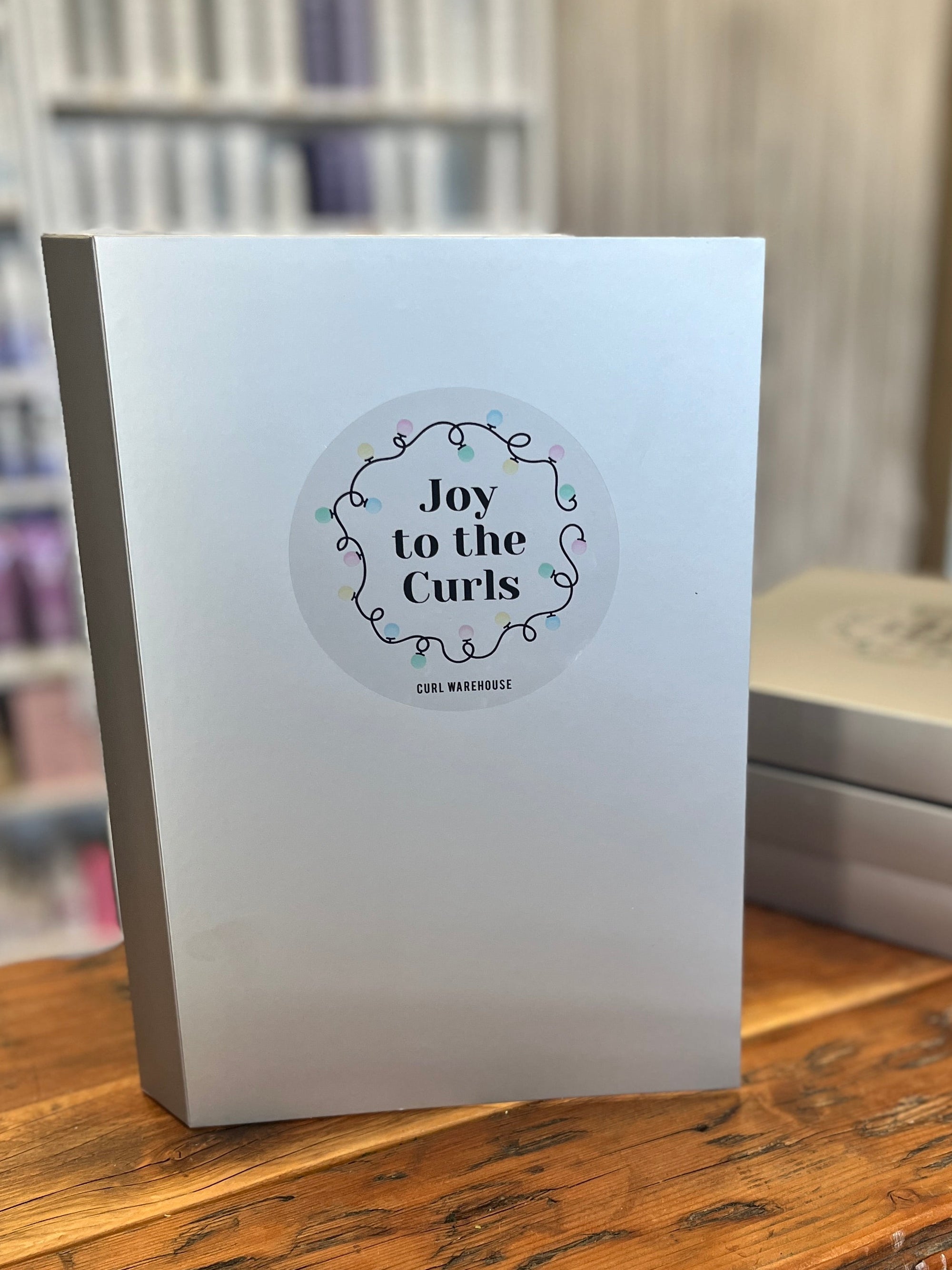 White advent calendar titled &#39;Joy to the Curls&#39; on a wooden surface with blurred shelves in the background
