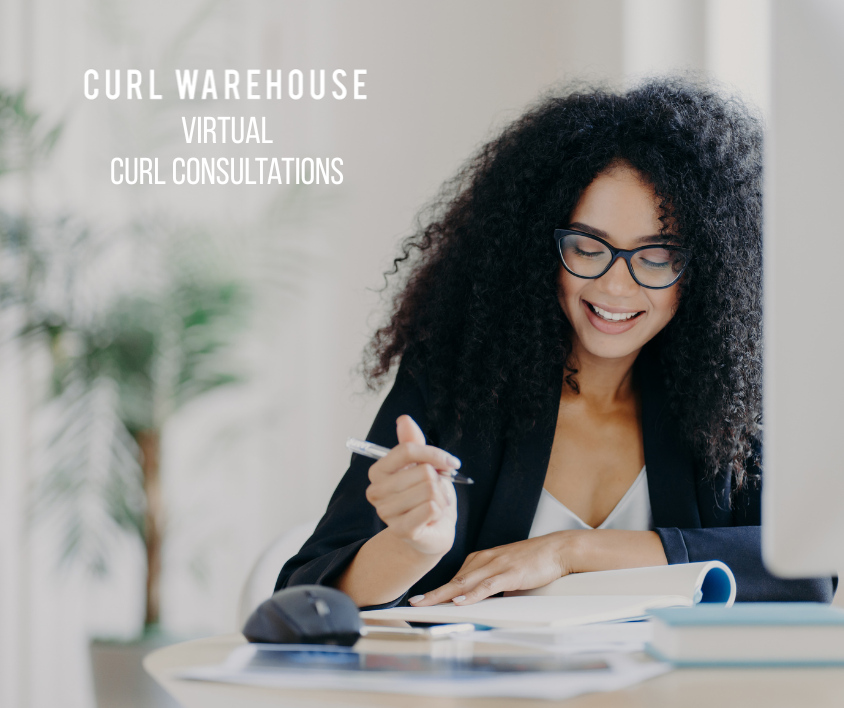 Woman with curly hair and glasses sitting at a desk, smiling and writing in a notebook.